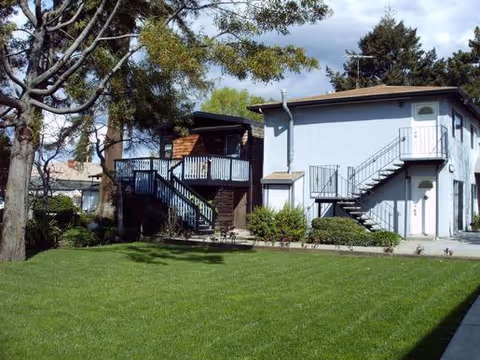 Exterior view of a two-story building with an outdoor staircase, surrounded by green grass, trees, and shrubs under a partly cloudy sky.