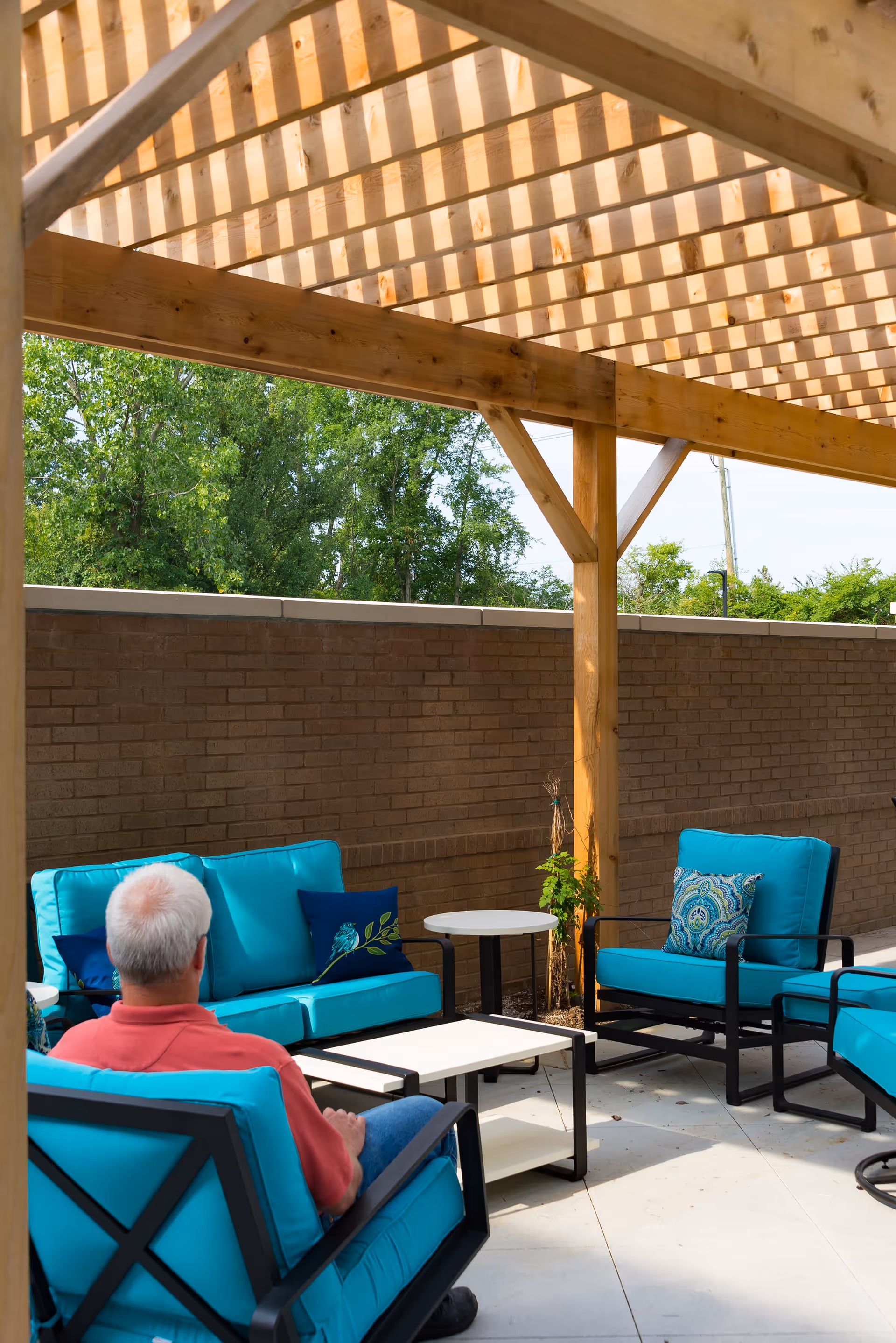 An outdoor patio area with a wooden pergola overhead, featuring turquoise cushioned chairs and sofas with decorative pillows. A man with gray hair wearing a red shirt and blue jeans is seated on one of the chairs, facing away from the camera. There are small tables placed between the seating arrangements, and a brick wall and green trees are visible in the background.