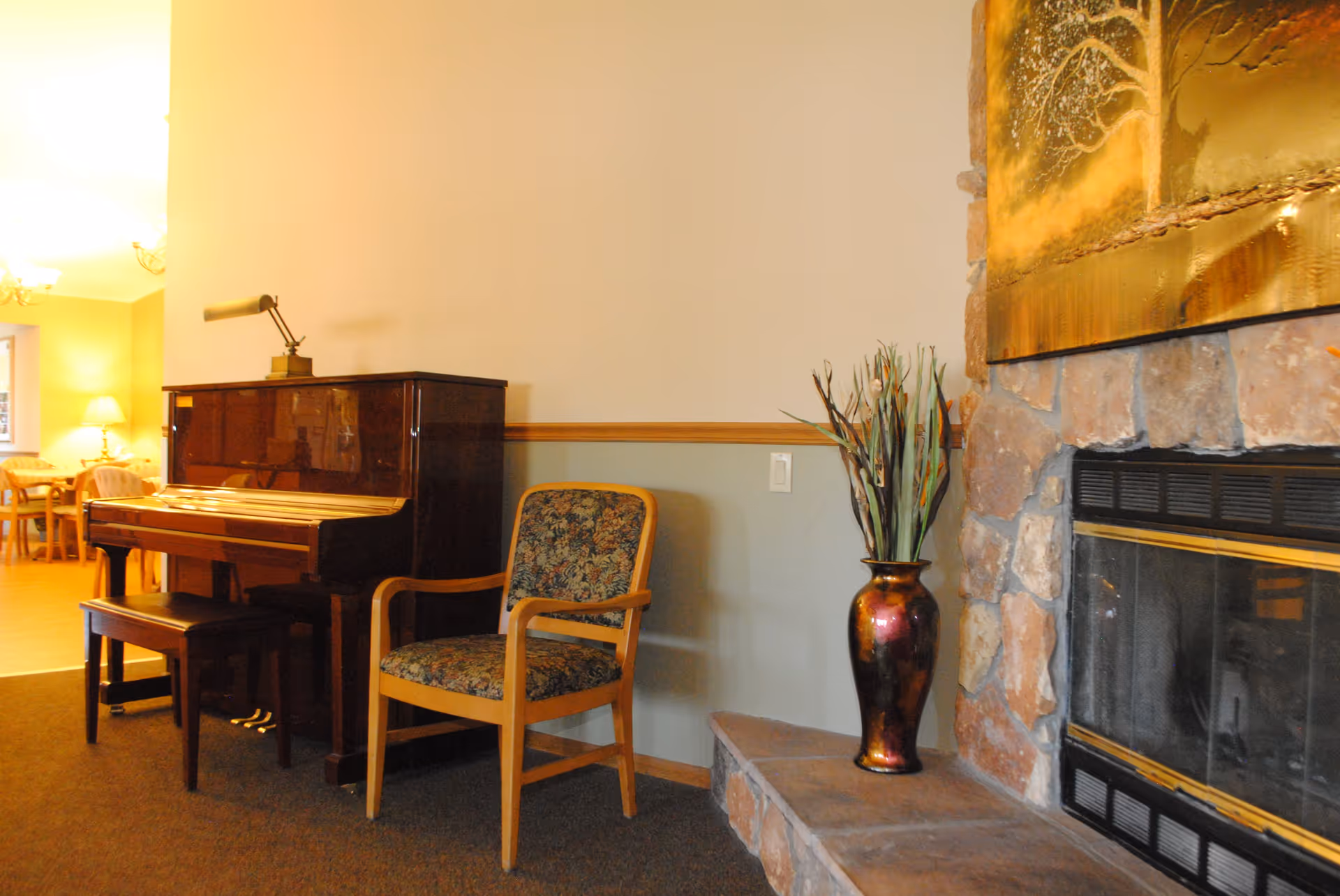 Interior view of a cozy living area featuring a dark wooden upright piano with a matching bench, a floral upholstered wooden armchair, a decorative vase with tall plants on a stone hearth next to a stone fireplace, and a glimpse of a dining area with tables and chairs in the background.
