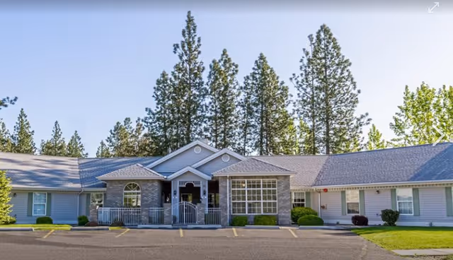 Exterior view of a single-story senior living facility building named Cornerstone Court, featuring a central entrance with a small porch, large windows, and surrounded by tall pine trees under a clear blue sky. The parking lot is visible in front of the building.