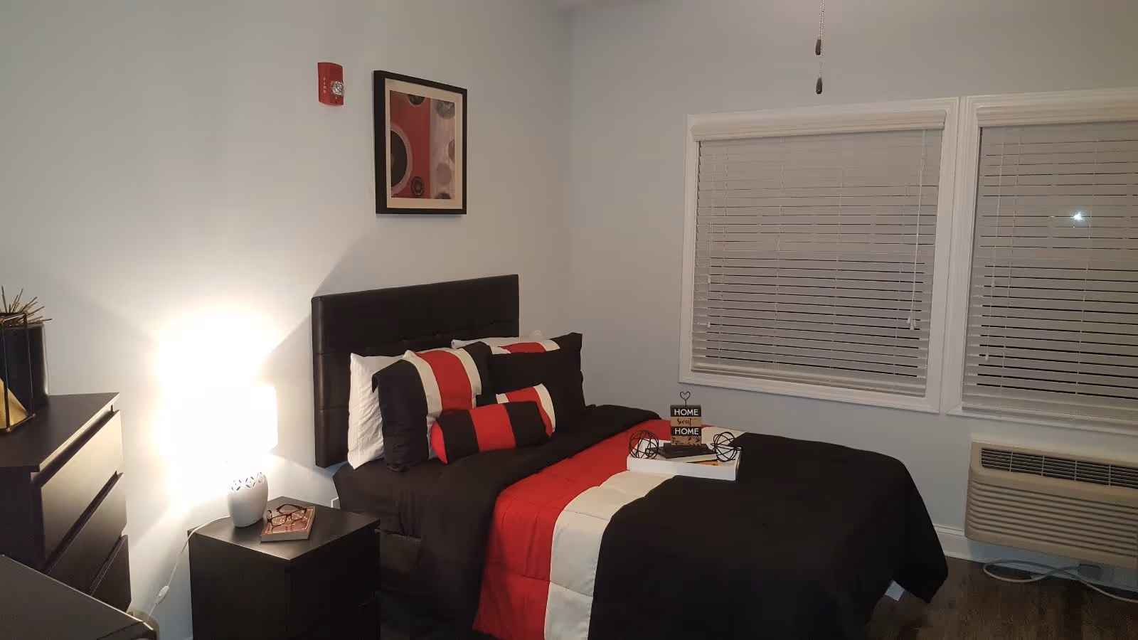 A neatly made bed with black, red, and white bedding in a senior care home bedroom. The room has a nightstand with a lamp and a small decorative item, a dresser, two windows with blinds, and an air conditioning unit below the windows. A framed abstract artwork hangs on the wall above the bed.