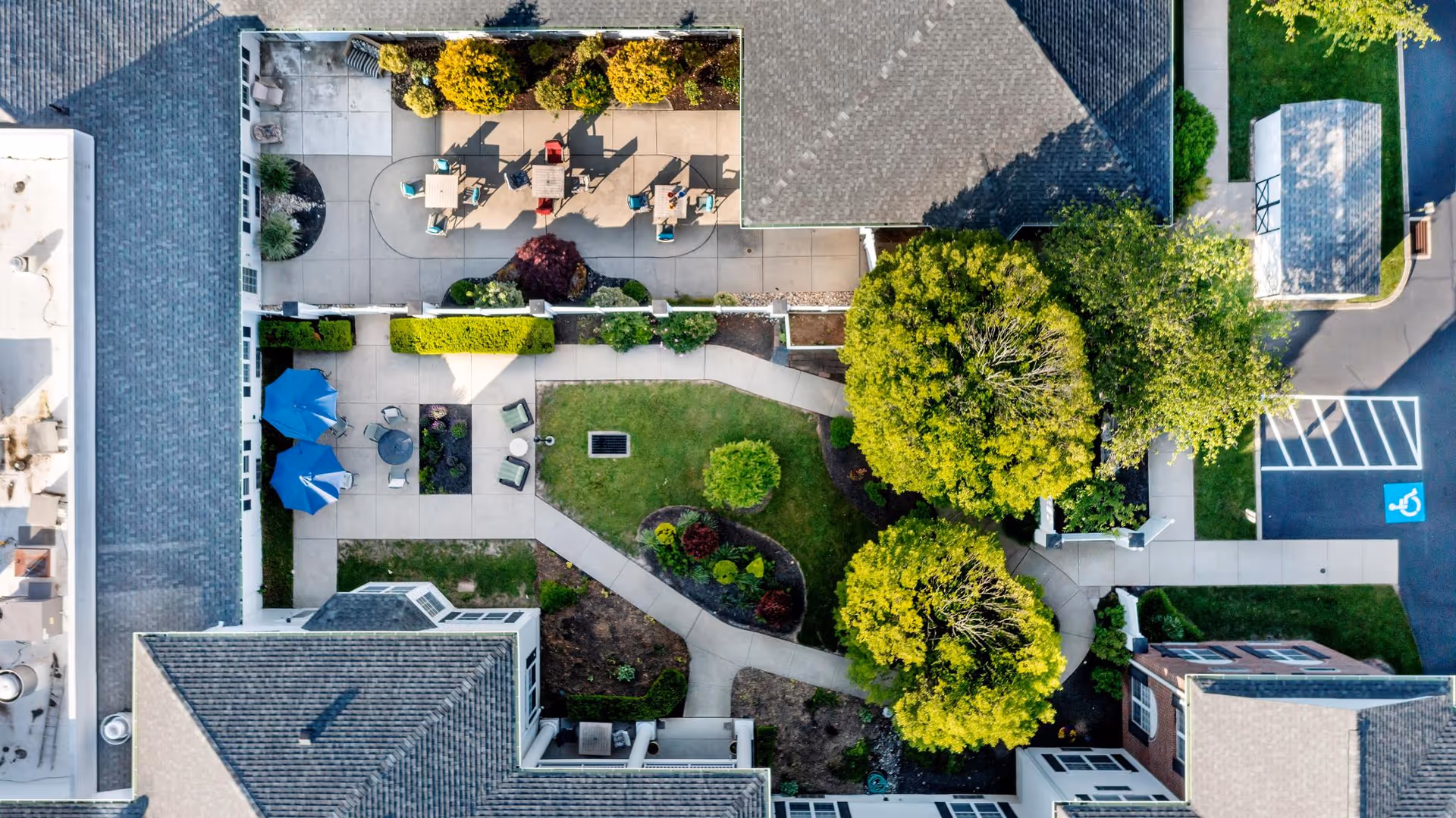 Aerial view of an outdoor courtyard area at Blue Bell Place featuring paved walkways, green lawns, trees, shrubs, patio tables with chairs, and blue umbrellas providing shade.
