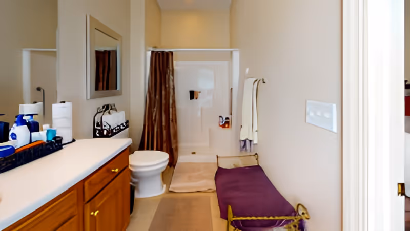Bathroom with a vanity and sink on the left, a toilet and shower with a curtain at the back, and a purple bench along the right wall.