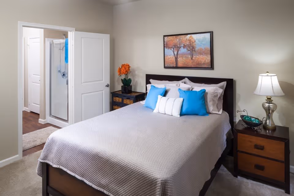 A neatly made bed with a light gray quilt and blue and white pillows in a bedroom. There is a nightstand with a lamp and decorative items on the right side of the bed. On the left side, there is a small cabinet with orange flowers in a vase. A framed painting of trees with autumn colors hangs on the wall above the bed. An open door reveals a bathroom with a glass shower enclosure and a blue towel hanging inside.