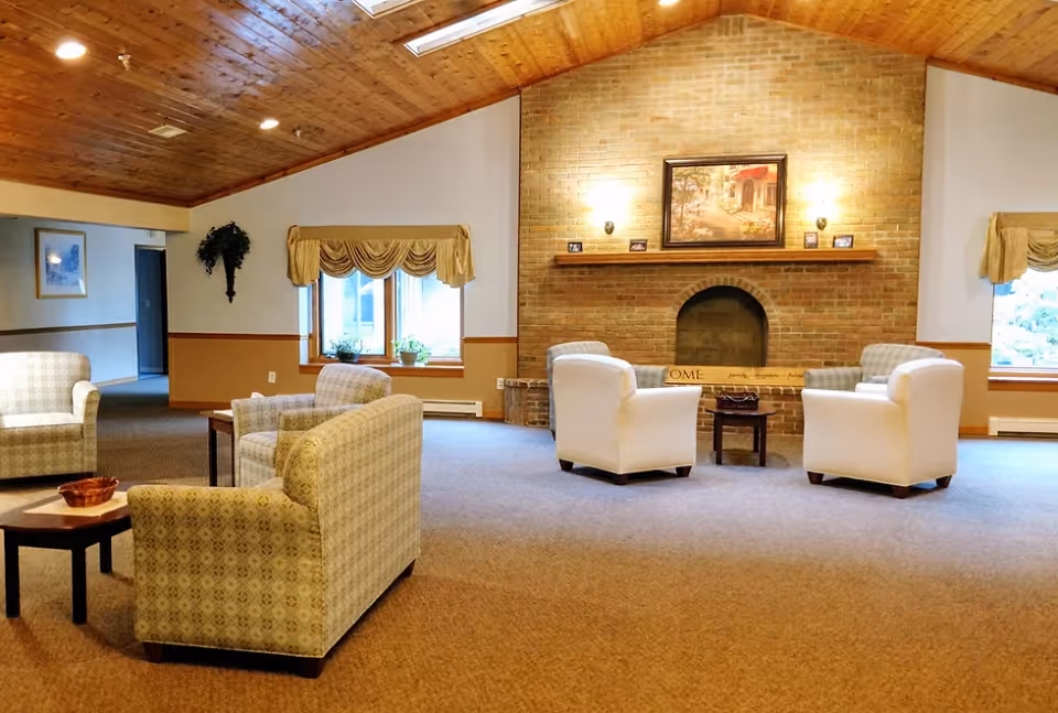 A spacious living room area with a high wooden ceiling and skylights. The room features a brick fireplace with a painting above it and two wall sconces on either side. There are four armchairs arranged around small tables, two white and two patterned. Two windows with valances let in natural light, and there is a plant hanging on the wall near a hallway.