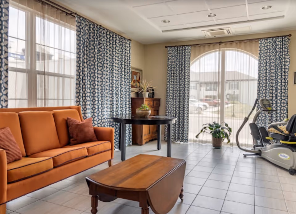A bright living room with an orange sofa adorned with two red pillows, a wooden coffee table, a round black table with a plant on it, and an exercise bike near large windows covered with sheer white and patterned blue curtains. Outside the windows, cars and buildings are visible.