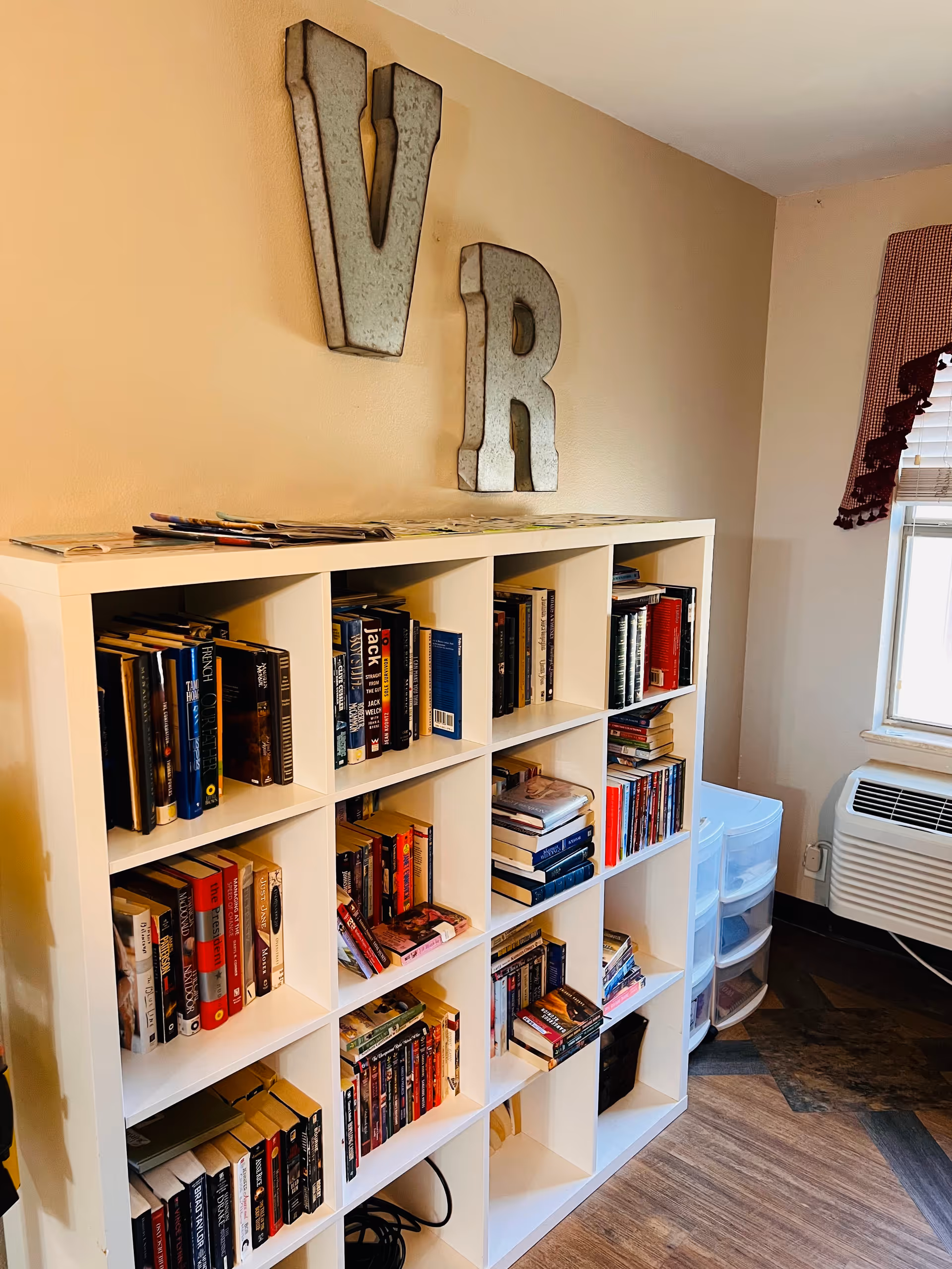 A white cubby bookshelf filled with various books is placed against a beige wall. Above the bookshelf, large metal letters 'V' and 'R' are mounted on the wall. To the right, there is a window with a red valance and a white air conditioning unit below it. The floor has a wood and tile pattern.