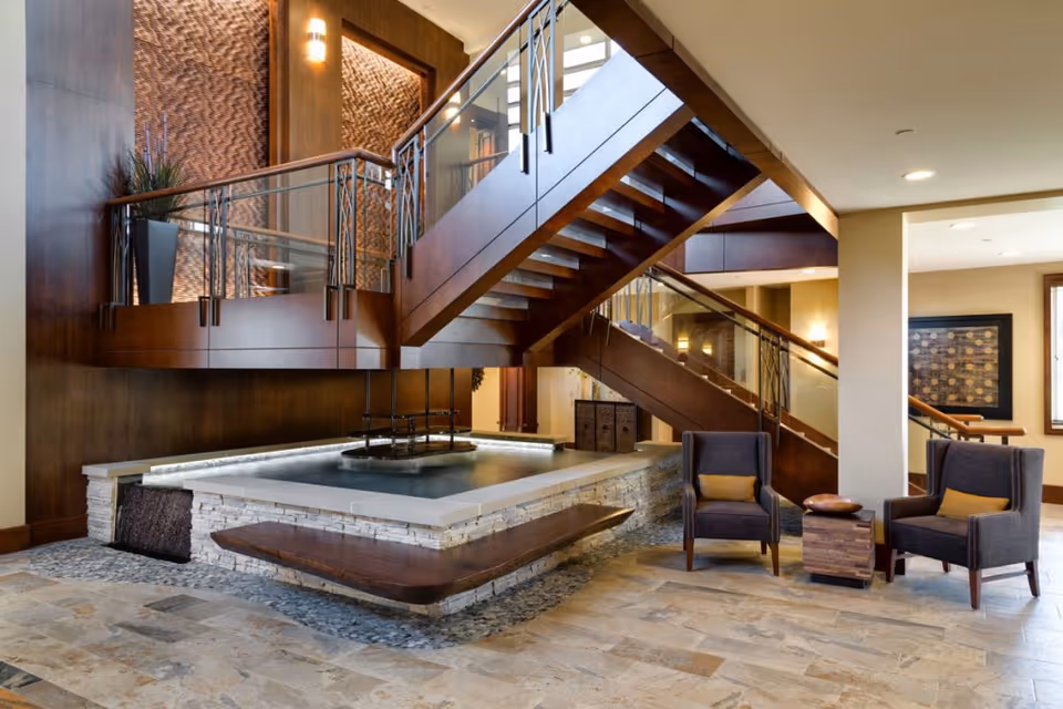 Interior view of a senior living facility lobby featuring a modern wooden staircase with glass railings, a water feature with a stone base, two gray armchairs with yellow cushions, a small wooden side table, and decorative wall art.