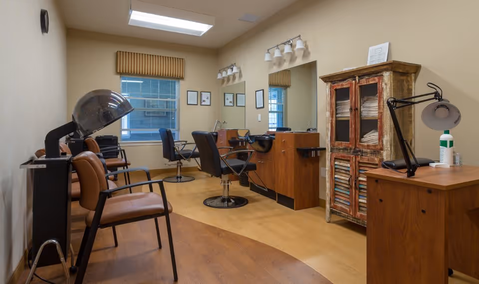 Interior view of a salon area in an assisted living facility featuring salon chairs, a hair dryer, mirrors with lights above, a wooden cabinet with towels, and a desk with a lamp and lotion bottle.