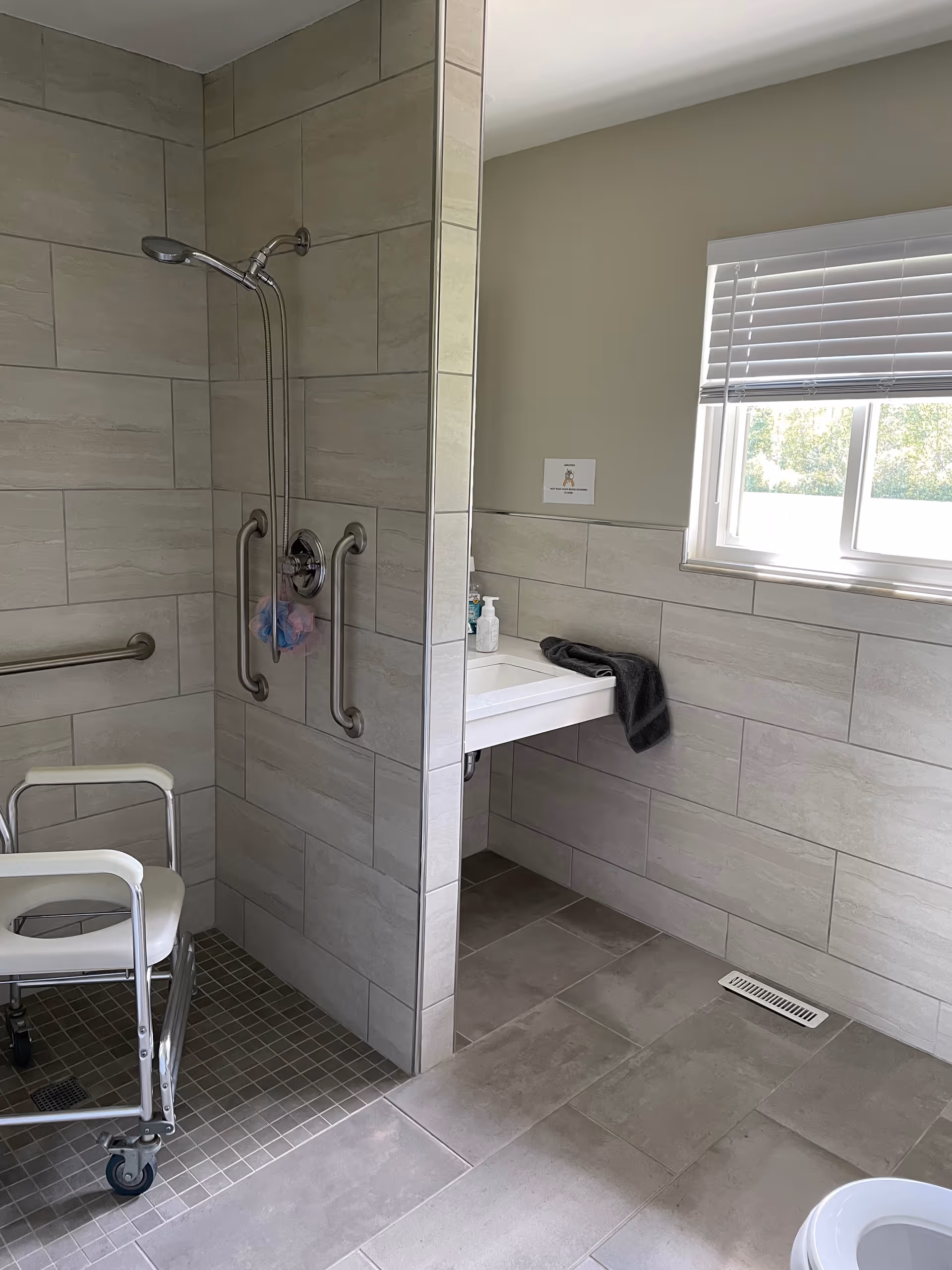 A bathroom with beige tiled walls and floor. It features a walk-in shower with a handheld showerhead, two grab bars, and a white shower chair with wheels. There is a white sink with a black towel on the counter, a soap dispenser, and a window with white blinds letting in natural light.