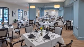 Bright, softly lit dining room with multiple tables set with white linens, napkins, and place settings surrounded by chairs.