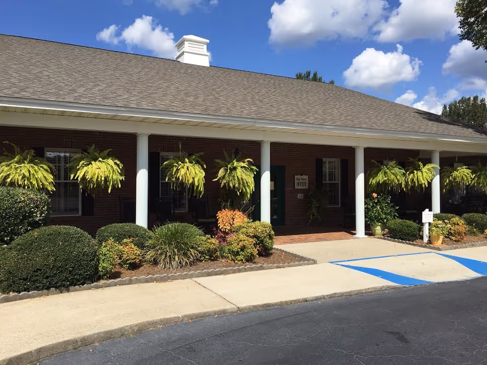 Exterior view of a single-story brick building with a covered porch supported by white columns. Several hanging green plants and well-maintained shrubs line the front of the building. A sign near the entrance reads 'Club House Office'. The sky is blue with scattered white clouds.