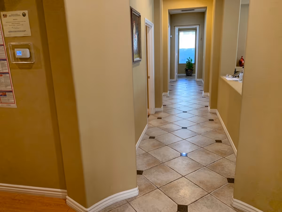 A long hallway with beige walls and tiled floor featuring a diamond pattern. There are doorways on both sides of the hallway, a framed picture on the left wall, and a potted plant at the far end near a window with blinds. A small counter with a red vase and some papers is visible on the right side.
