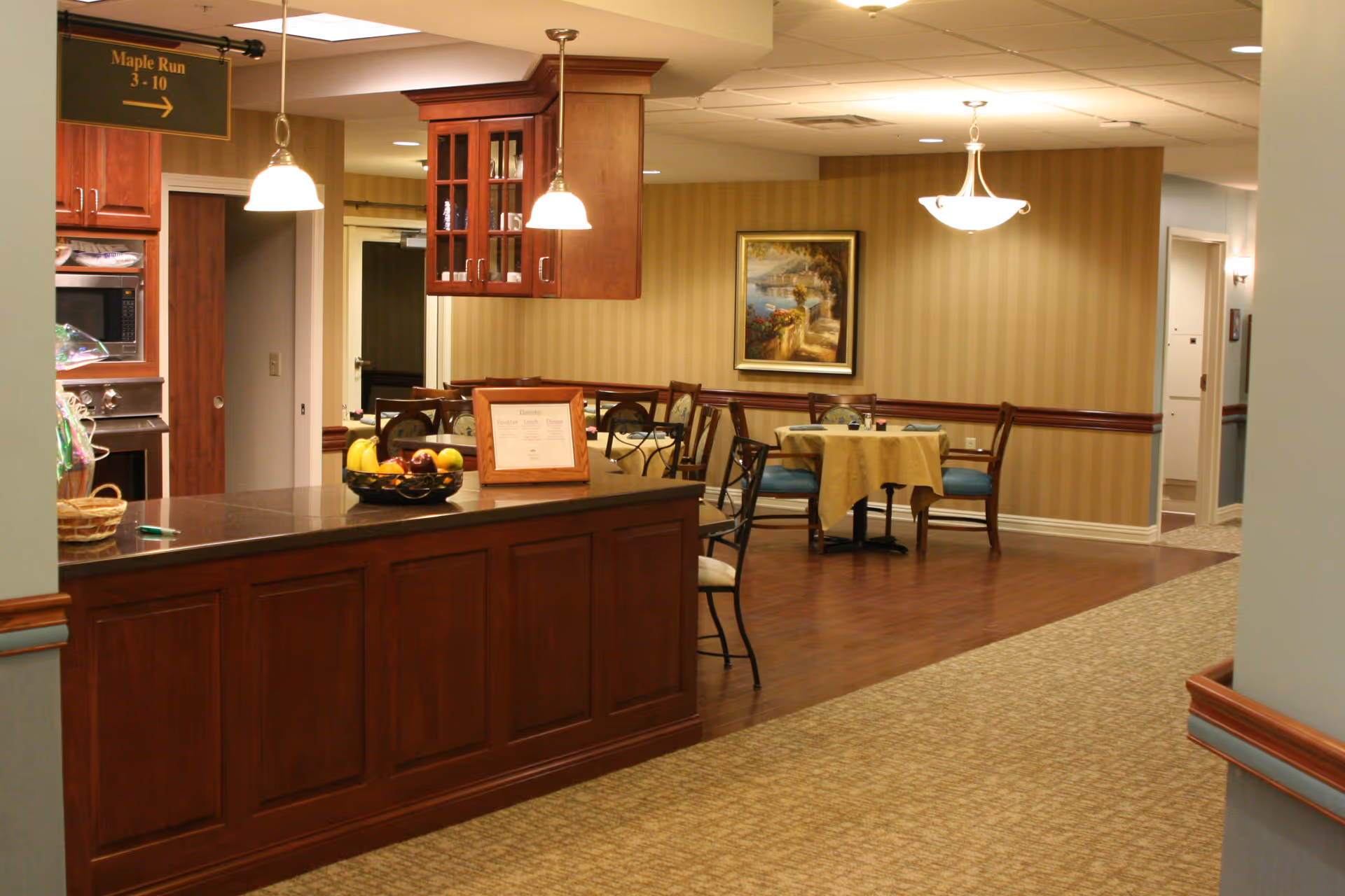 Interior view of a senior living facility dining area with wooden cabinetry, a countertop with a fruit basket and framed menu, several tables with chairs, warm lighting, and a painting on a striped wallpapered wall.