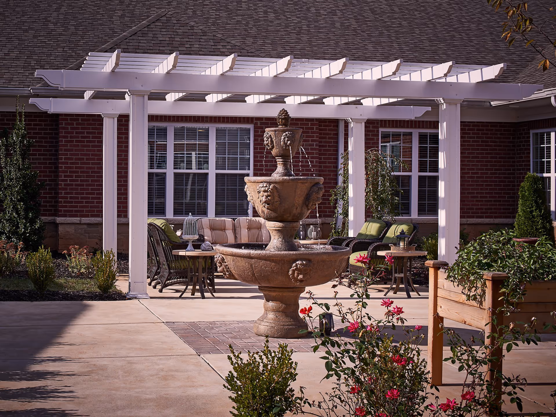 Outdoor patio area with a large stone fountain in the center, surrounded by flowering plants and greenery. Behind the fountain is a white pergola with cushioned outdoor seating and tables. The background shows a brick building with multiple windows.