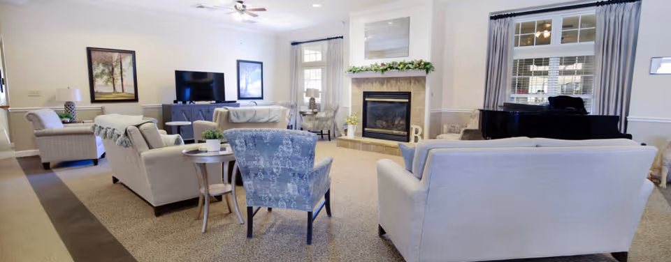 A spacious and well-lit living room area in a senior living facility featuring multiple sofas and armchairs arranged around a fireplace. There is a flat-screen TV on a stand, a grand piano near a window with curtains, and framed artwork on the walls. The room has a neutral color palette with beige carpeting and light-colored walls.