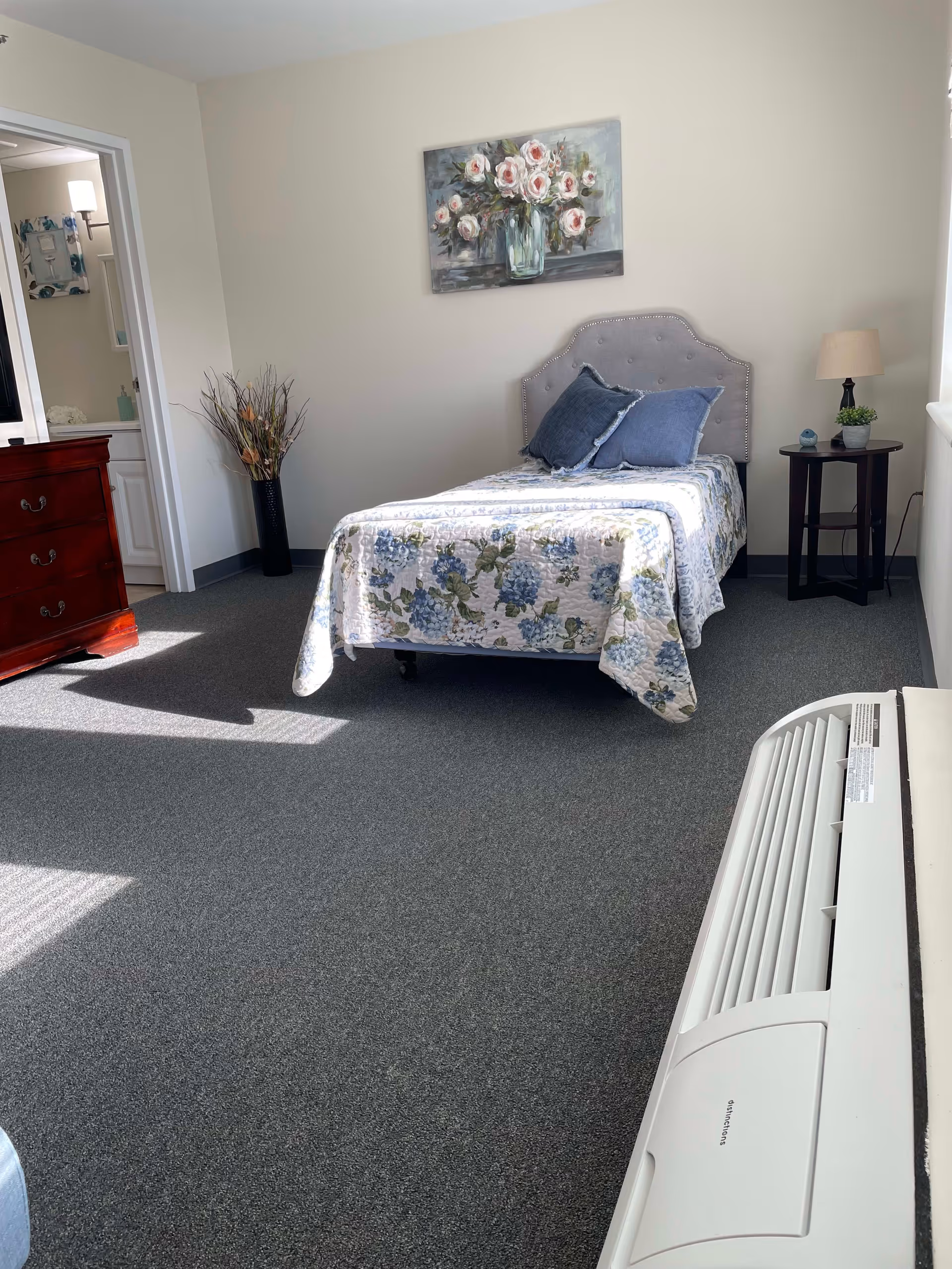 A small bedroom with a single bed covered in a floral quilt and two blue pillows. There is a nightstand with a lamp and a small plant next to the bed. A painting of flowers hangs on the wall above the bed. A dresser is visible to the left, and an open doorway leads to a bathroom. The floor is carpeted and there is a white air conditioning unit in the foreground.