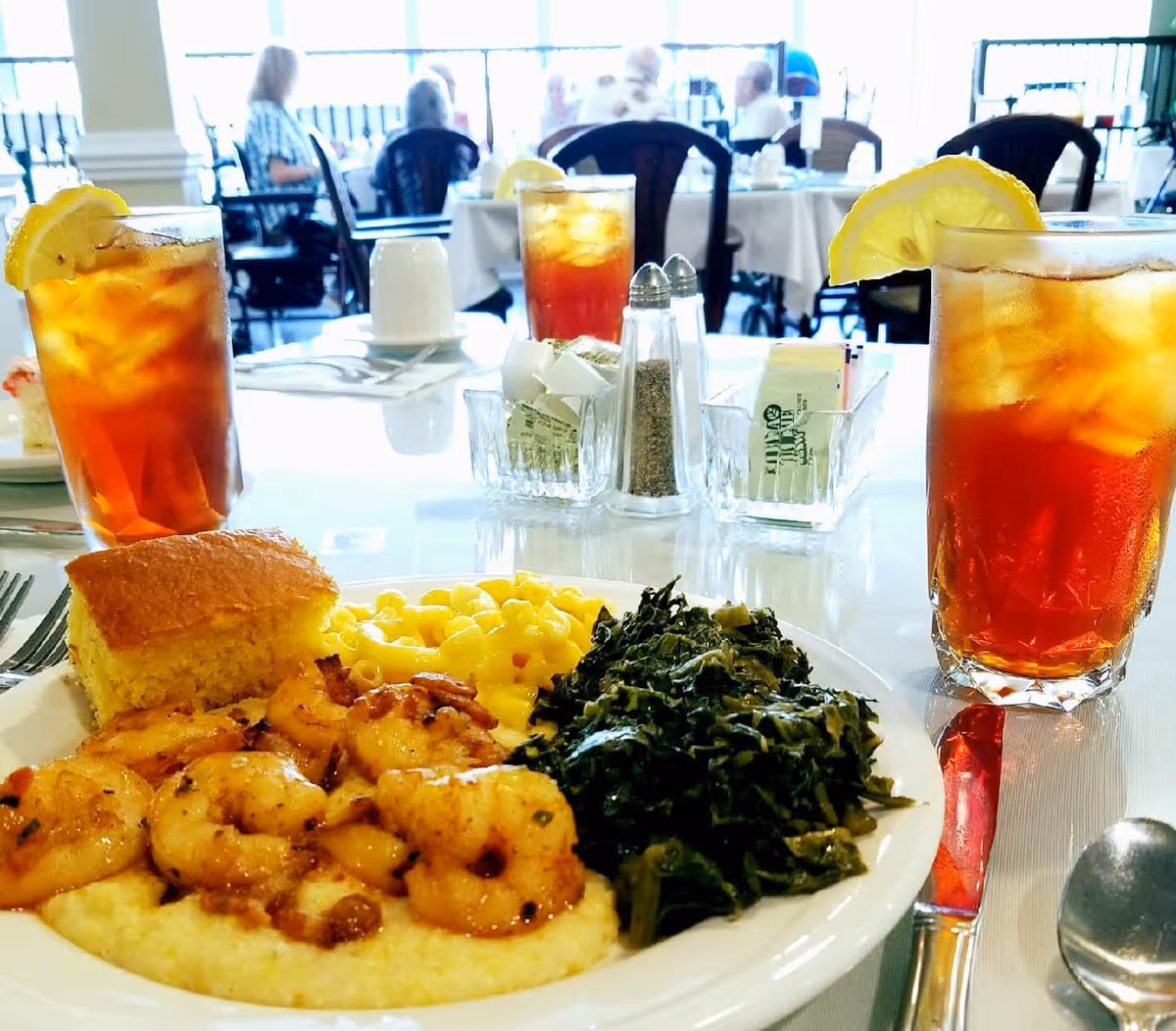 A close-up of a plate with shrimp on grits, macaroni and cheese, cooked greens, and a piece of cornbread on a dining table. Two glasses of iced tea with lemon wedges are also on the table. In the background, several people are seated at tables in a dining room with large windows.
