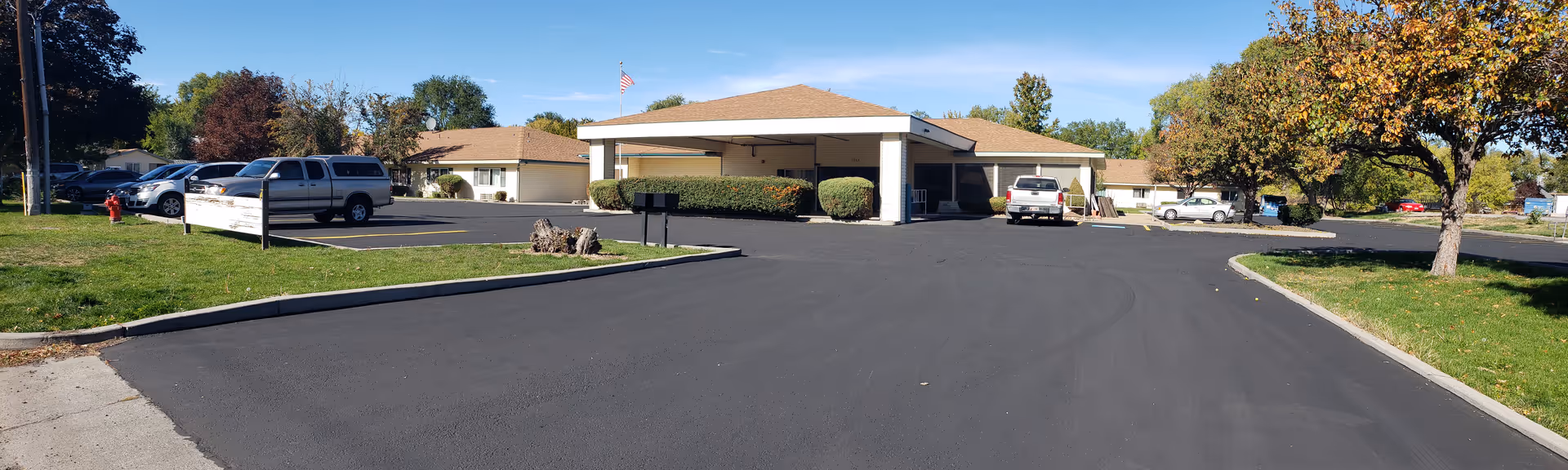 Single-story assisted living building with a covered entrance and parking lot, several cars, trees and an American flag under a clear blue sky.