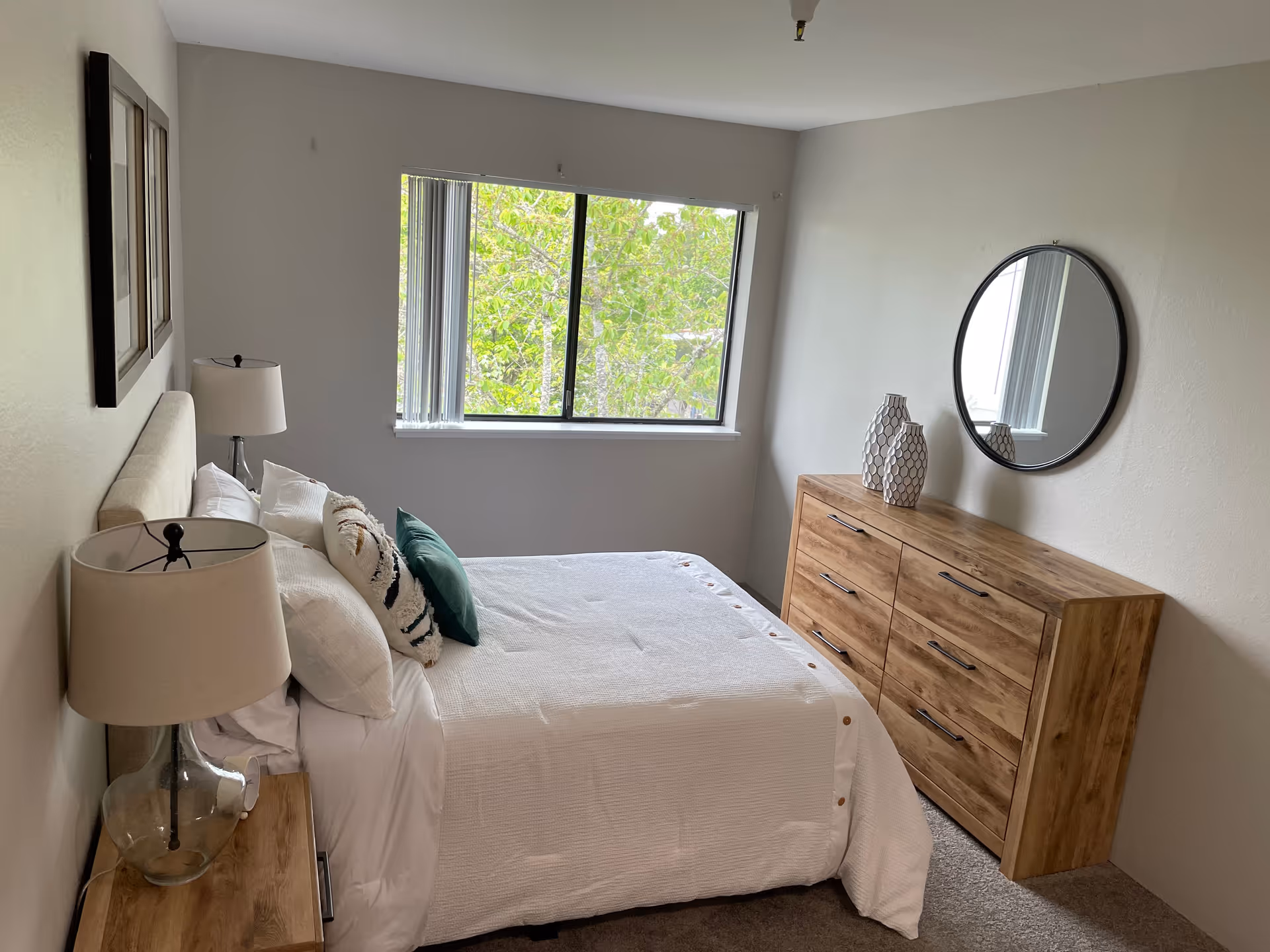 A neatly made bedroom with a bed, two lamps, a dresser, round mirror, and a window.