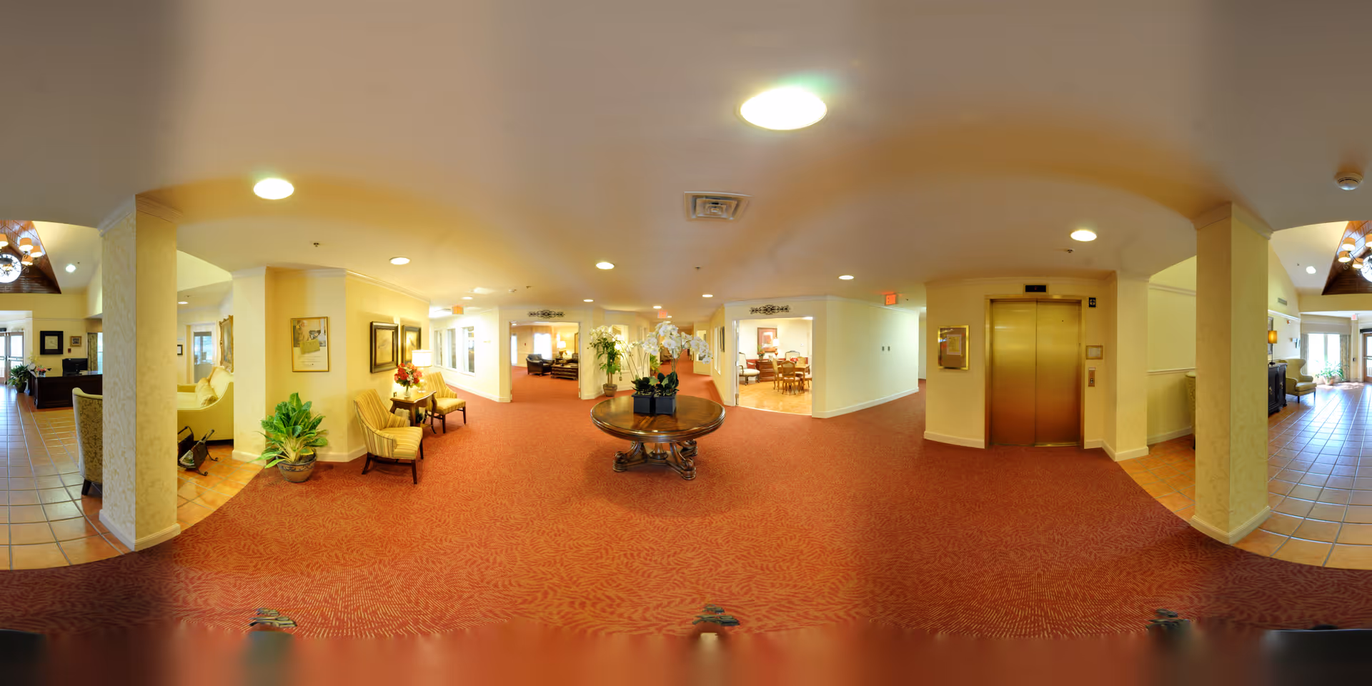 Wide panoramic view of a senior living facility interior hallway with red patterned carpet, a round wooden table with white orchids in the center, beige walls, and ceiling lights. There are seating areas with chairs and plants on the left side, an elevator with golden doors on the right, and doorways leading to other rooms including a dining area in the background.