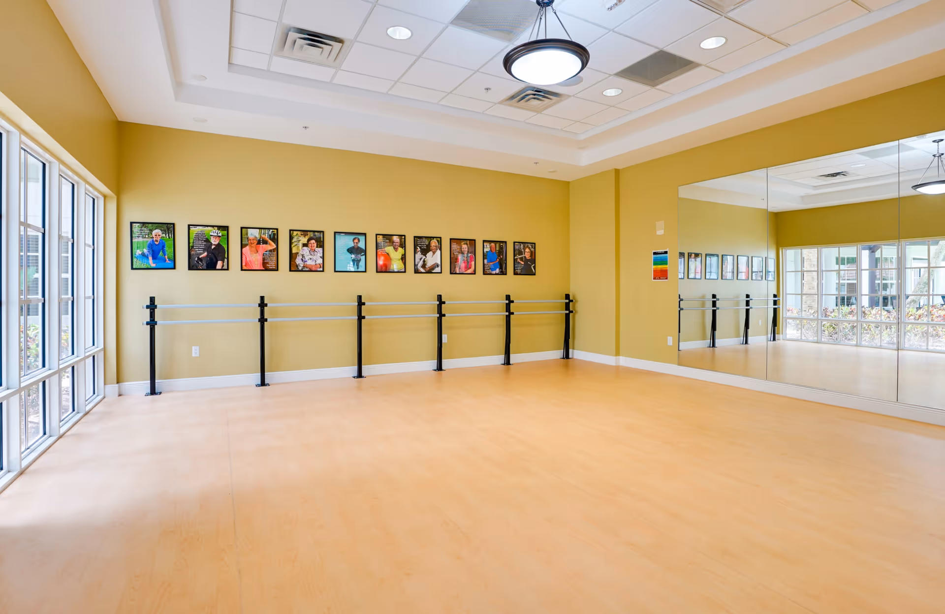 A spacious exercise or dance room with light wooden flooring, yellow walls, and a large mirror covering one wall. There are ballet barres mounted on the wall beneath framed photos of people. Large windows on one side let in natural light and provide a view of the outdoor area.