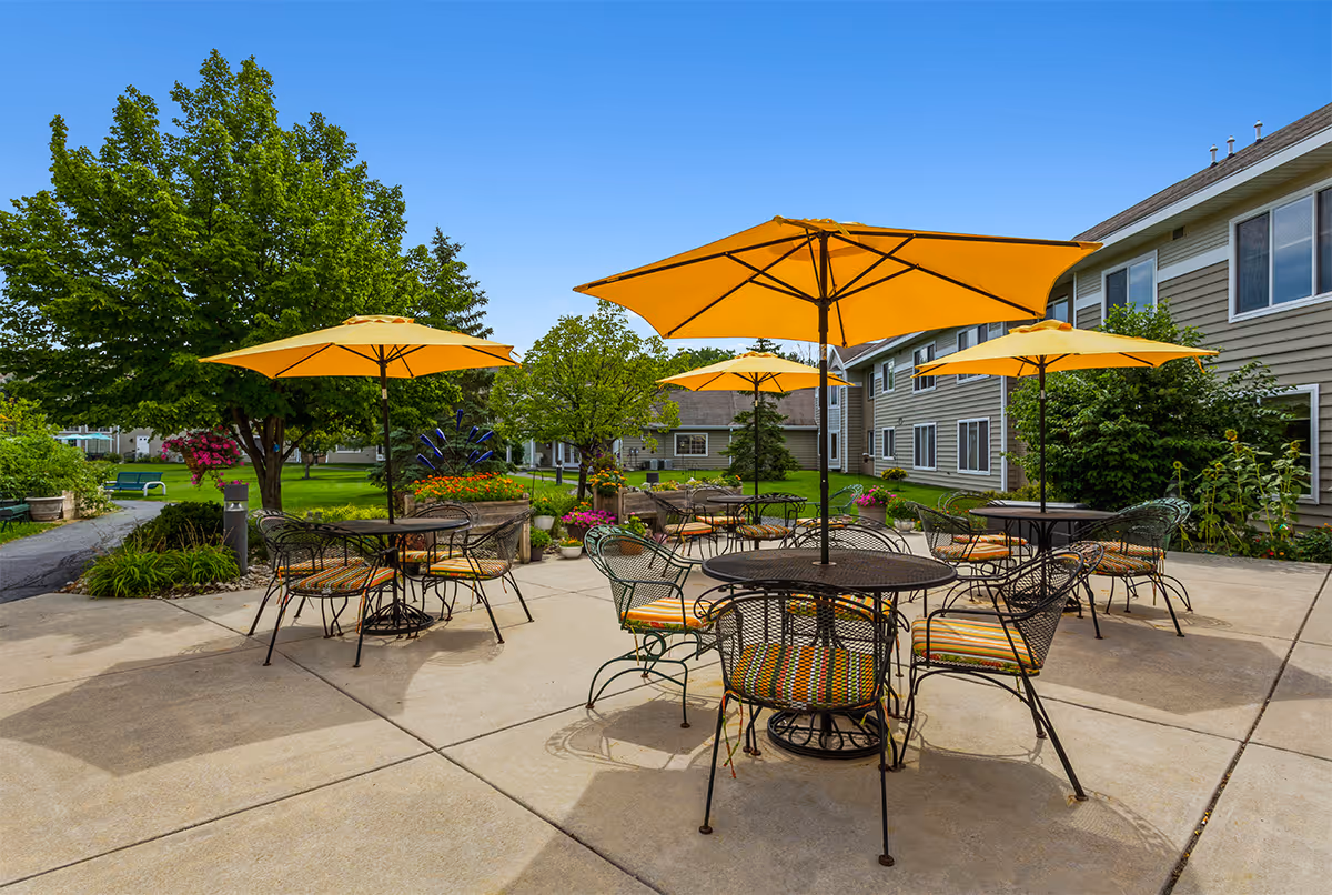 Outdoor patio area with several round metal tables and chairs, each table shaded by a large yellow umbrella. The patio is surrounded by green grass, trees, and flowering plants, with a two-story building in the background under a clear blue sky.
