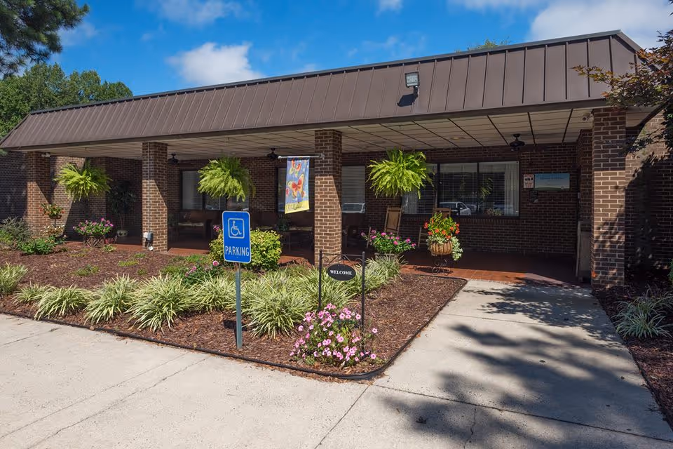 Covered front entrance of a brick building with hanging ferns, potted flowers, a 'Welcome' sign, and a handicapped parking sign.