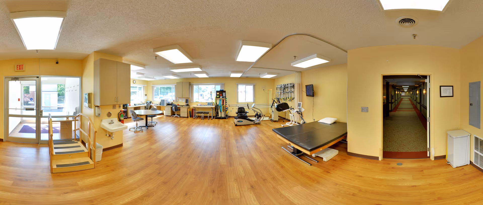 Well-lit interior therapy and activity room with exercise equipment, a treatment table, seating, and an entrance leading to a hallway.