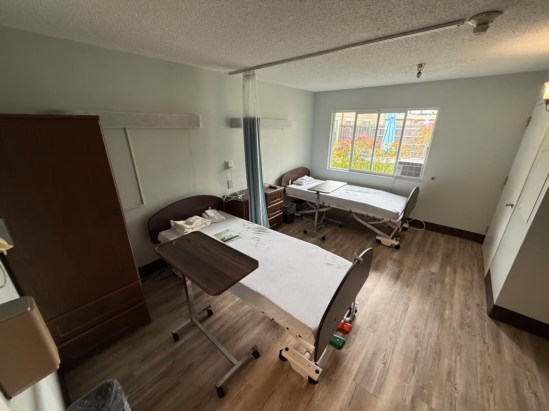 A bright shared patient bedroom with two adjustable hospital beds, overbed tables, a privacy curtain, wardrobe and a window overlooking landscaping.