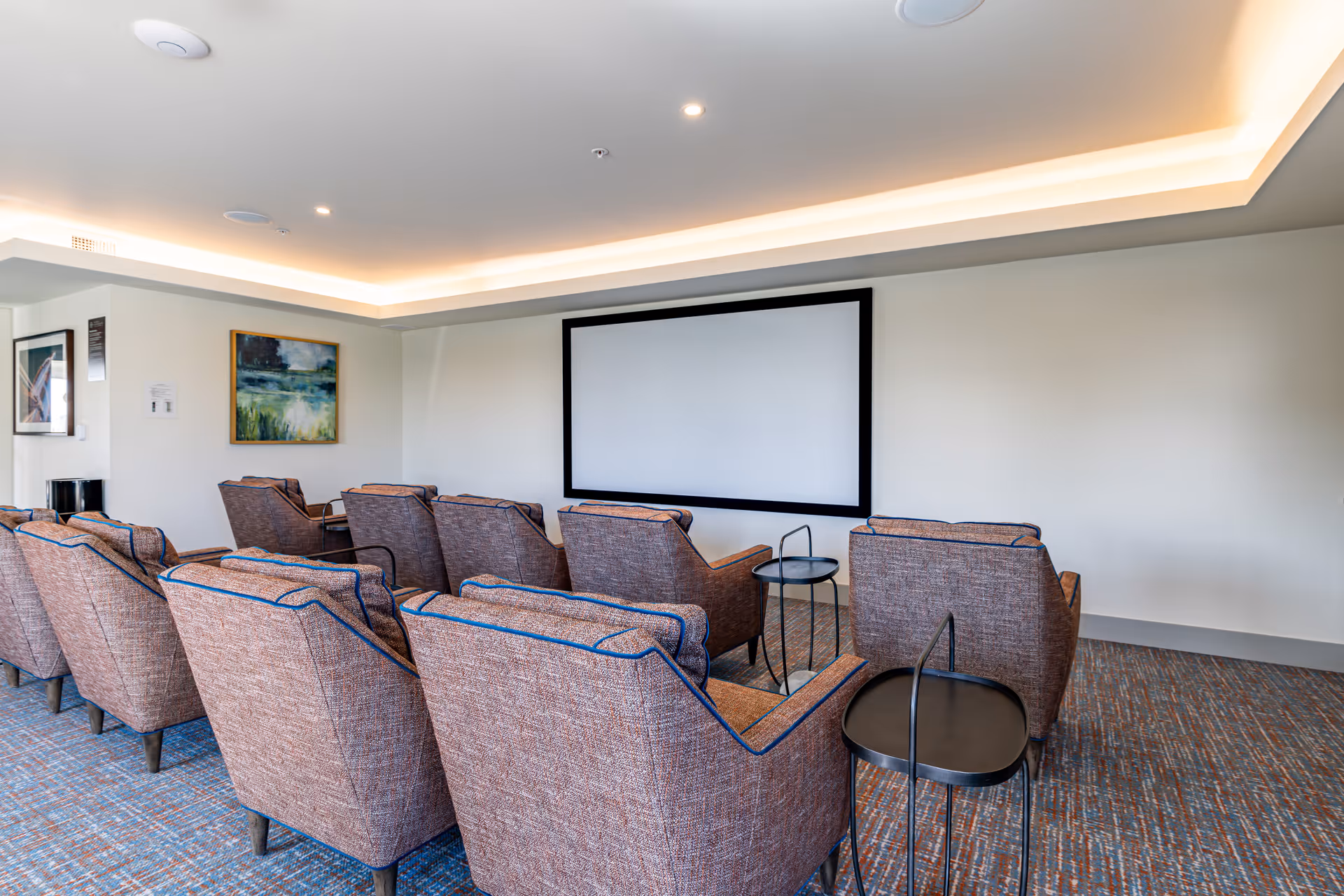 Small screening room with rows of upholstered armchairs facing a blank projection screen and side tables.