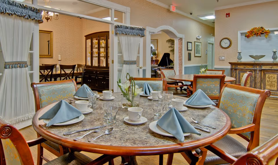 A dining room in a senior living facility with a round table set for six people. Each place setting includes a plate with a folded blue napkin, a cup and saucer, a glass, and silverware. The chairs have wooden frames with patterned upholstery. In the background, there are more tables and chairs, a wooden cabinet, and decorative elements such as curtains, framed pictures, and a clock on the wall.