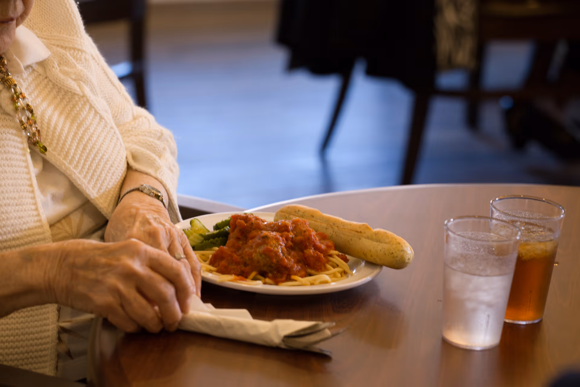 An elderly person wearing a white sweater and a beaded necklace is sitting at a wooden table with a plate of spaghetti topped with tomato sauce and meatballs, a breadstick, and some green vegetables. There are two glasses on the table, one with water and the other with iced tea.