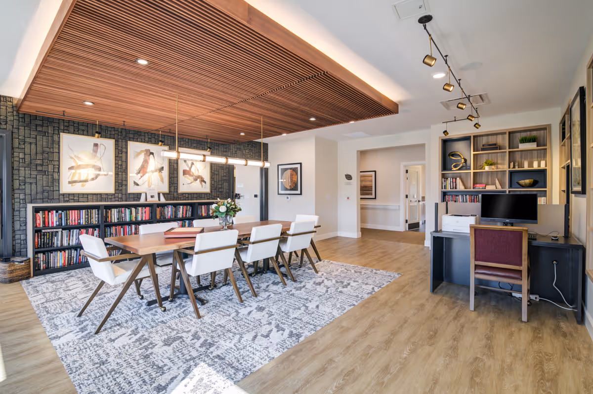Bright modern communal room with a long wooden table and white chairs on a patterned rug, bookshelves and artwork along one wall, and a desk with a computer to the right.