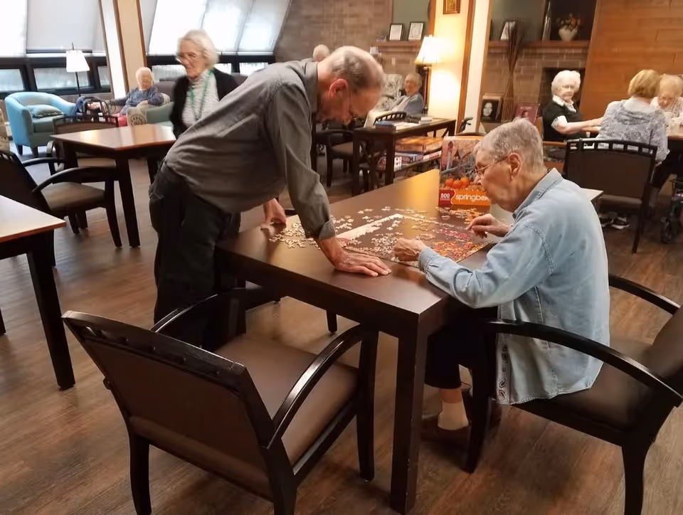Elderly residents at Wellhaven Senior Living are engaged in assembling a jigsaw puzzle on a wooden table in a common room with several other tables and chairs. The room has large windows, wooden flooring, and a cozy atmosphere with shelves and framed pictures on the walls.
