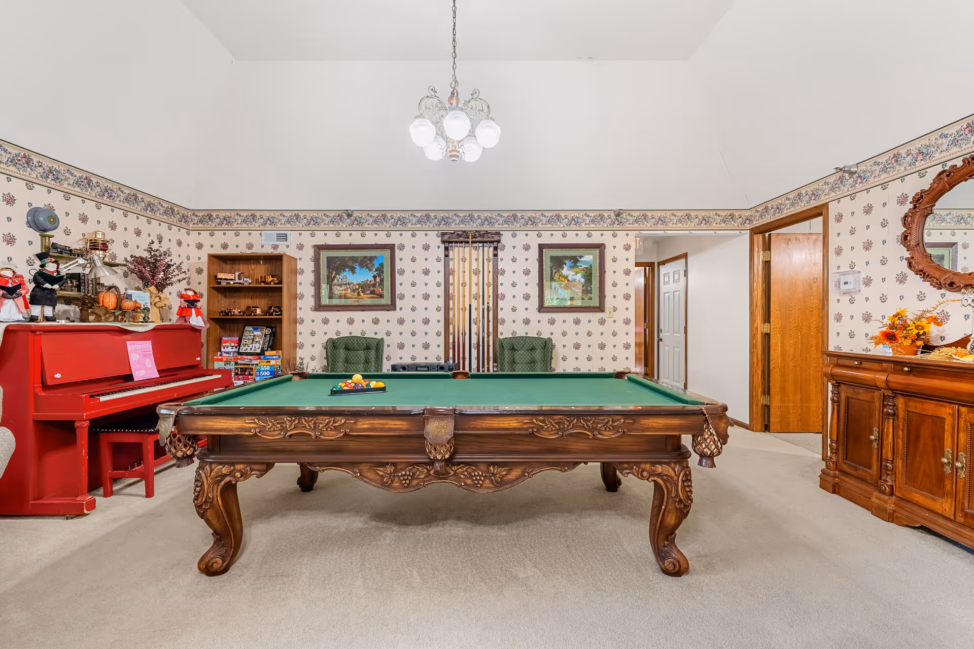 A recreational room with a large ornate wooden pool table in the center, set up with billiard balls and cues. The room features floral wallpaper with a border near the ceiling, two green upholstered chairs against the back wall, and framed paintings. To the left, there is a bright red piano with decorative items on top, and a wooden bookshelf filled with games and puzzles. On the right side, there is a wooden sideboard with a decorative mirror above it and a floral arrangement. The room is carpeted and well-lit by a chandelier hanging from the ceiling.