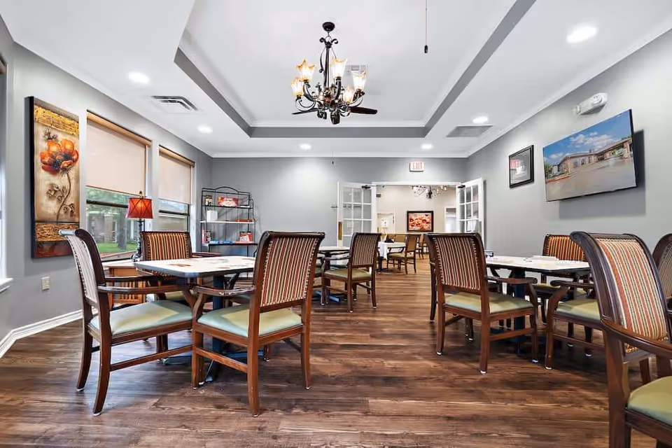 A bright dining room with several wooden tables and upholstered chairs, a chandelier, wall art, and a TV.