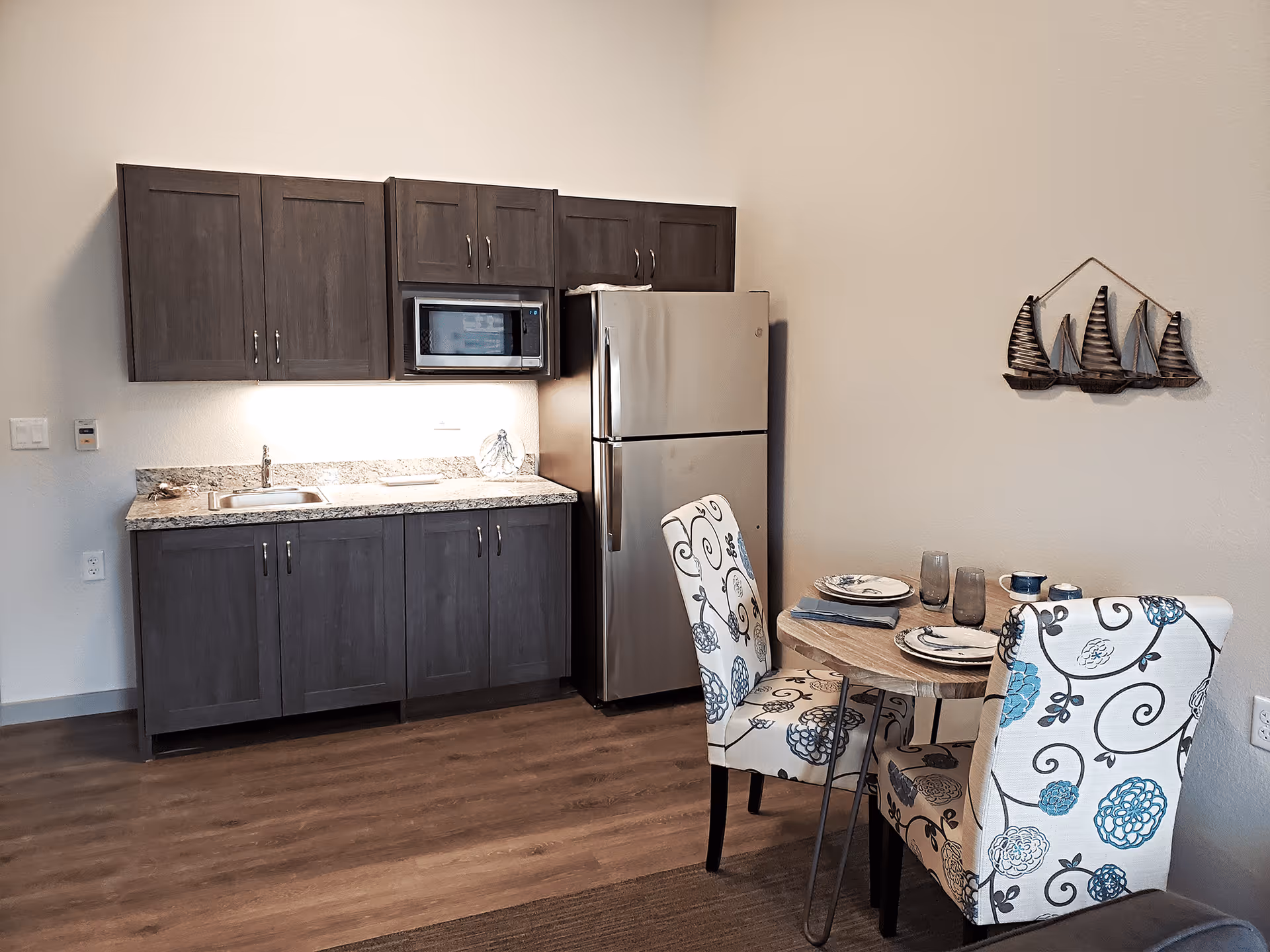 Small kitchenette with gray cabinets, stainless refrigerator and microwave, and a round dining table with two patterned chairs.