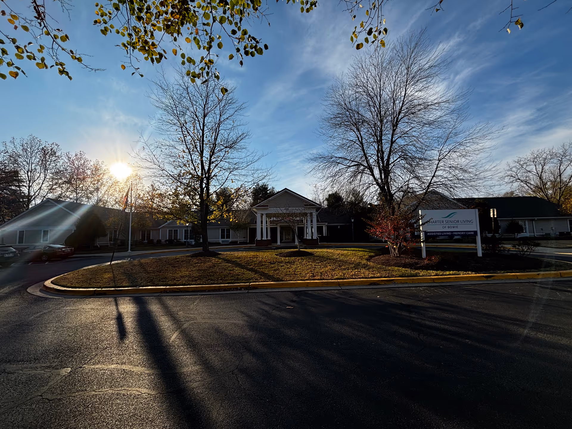 Exterior view of Charter Senior Living of Bowie building with a driveway and a lawn area with trees in front. The sun is low in the sky, casting long shadows, and there is a sign on the right side indicating the facility name and services.