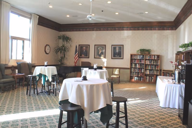 Bright communal dining/lounge room with round tables draped in white cloths, stools, bookshelves, framed portraits, and seating near windows.