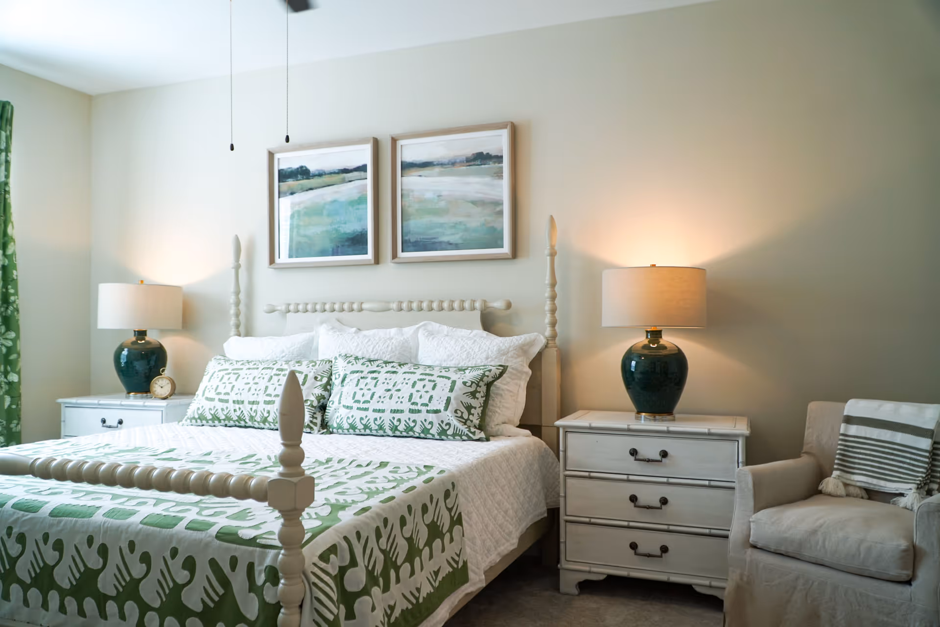 A cozy bedroom featuring a white wooden bed with green and white patterned bedding and pillows. On either side of the bed are white nightstands with dark green lamps. Above the bed are two framed landscape paintings. To the right is a beige armchair with a striped throw blanket draped over it. The room has light-colored walls and a window with green curtains on the left.