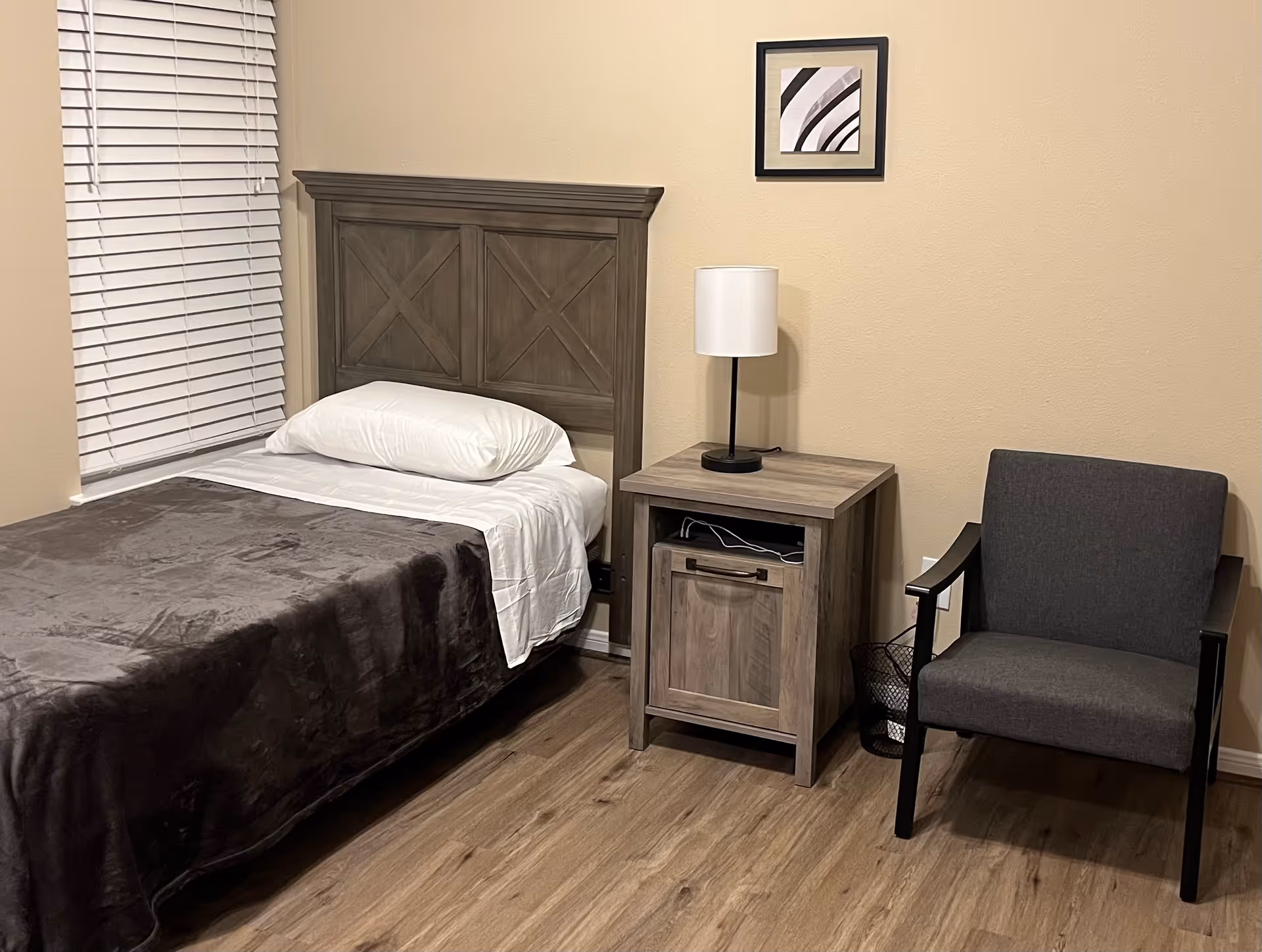 A tidy bedroom with a single bed against a wooden headboard, a nightstand with a lamp, and a gray armchair.