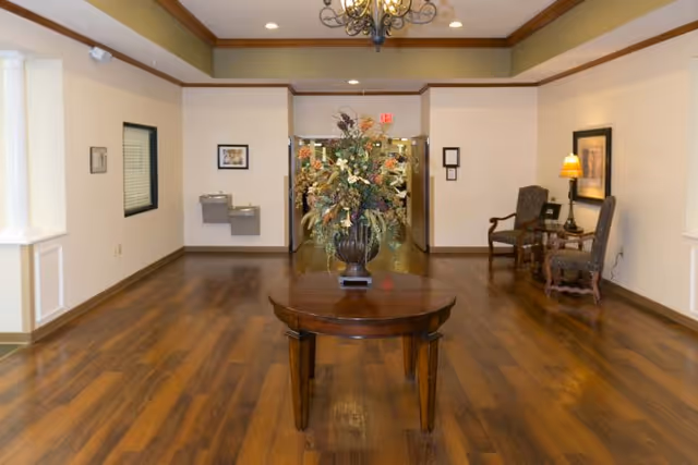 Interior view of a hallway or lobby area with wooden flooring, a round wooden table with a large floral arrangement in the center, two upholstered chairs with a small table and lamp on the right side, water fountains on the left wall, and a chandelier hanging from the ceiling.