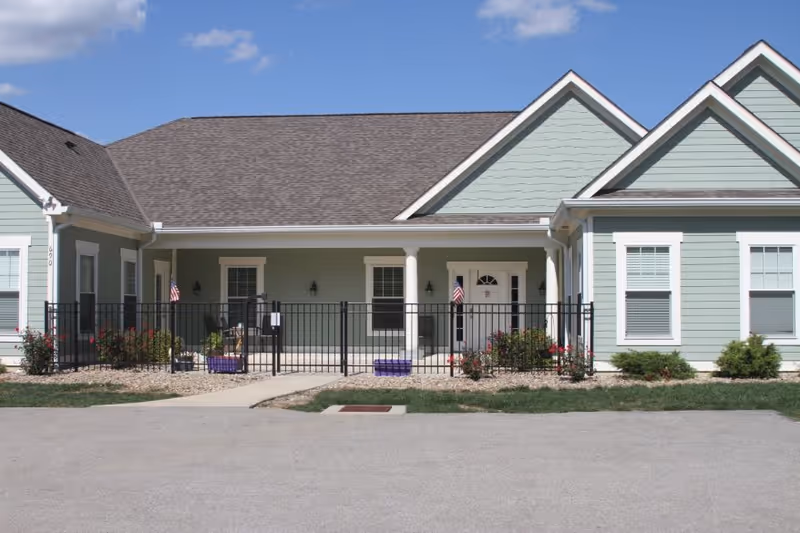 Exterior view of a single-story senior living facility building with light green siding, white trim, and a gray shingled roof. The entrance area is fenced with black metal fencing and has small garden beds with flowers and shrubs. Two small American flags are displayed near the entrance door under a covered porch. The sky is clear and blue with a few clouds.