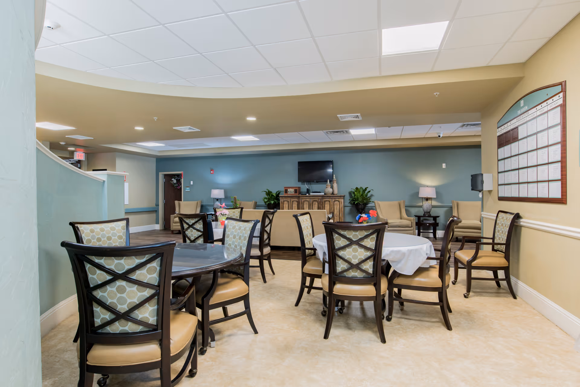 A bright communal dining and seating area with multiple round tables and chairs and a TV against a teal accent wall.