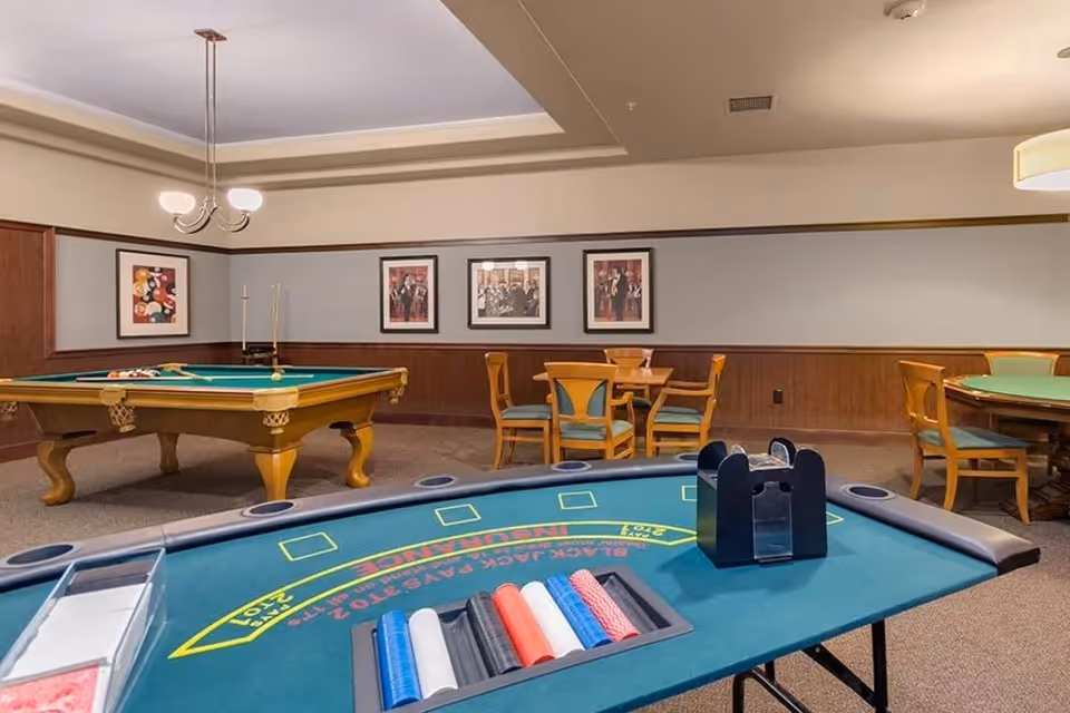 A game room featuring a poker table with chips and card holders in the foreground, a pool table with cues and balls in the background, and several wooden chairs and tables along the walls. The room has framed artwork on the walls and a ceiling light fixture.
