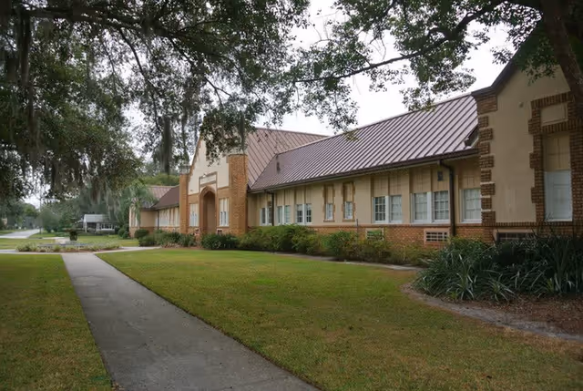 Exterior view of a single-story brick building with a metal roof, surrounded by grass and trees, with a concrete walkway leading up to the entrance.