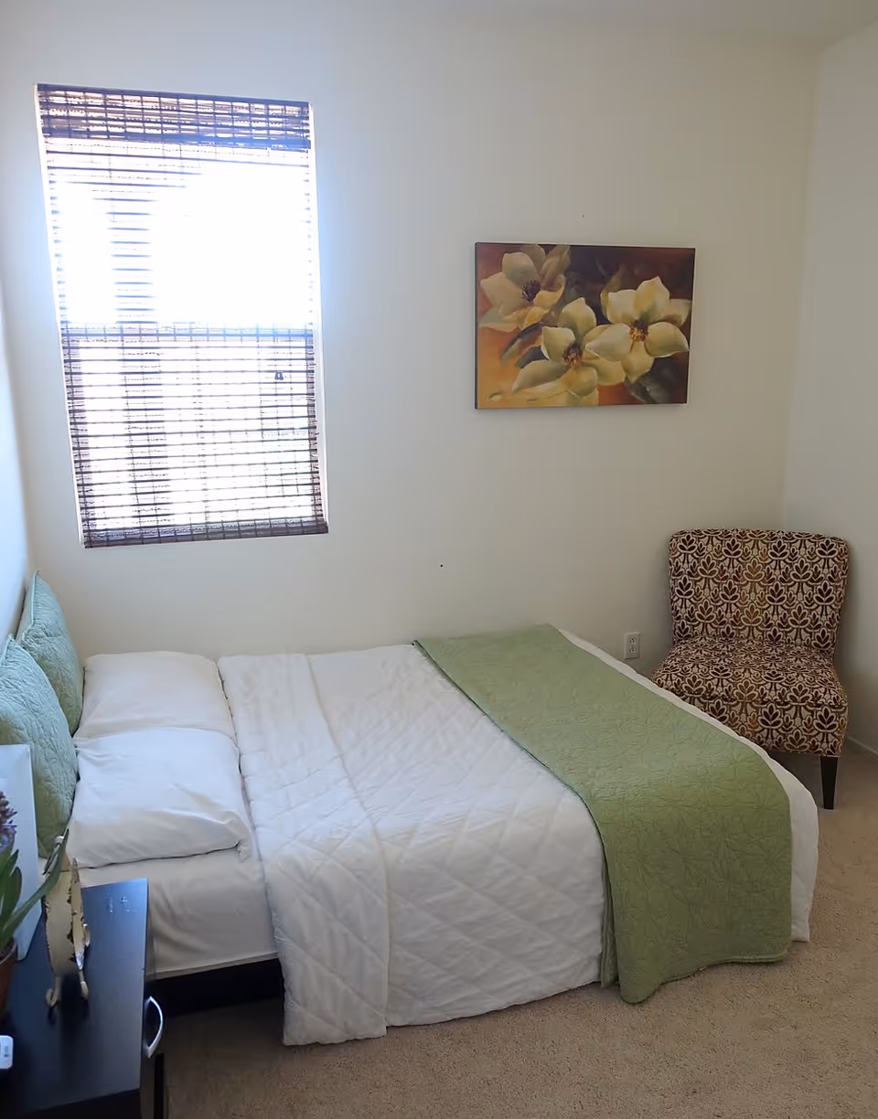 A small bedroom with a neatly made bed featuring white bedding and a green quilted blanket. There are two green pillows against the headboard. A window with a bamboo blind lets in bright light. On the wall above the bed is a painting of yellow flowers. A patterned upholstered chair is placed in the corner of the room, and a small black nightstand with a plant and decorative items is beside the bed.