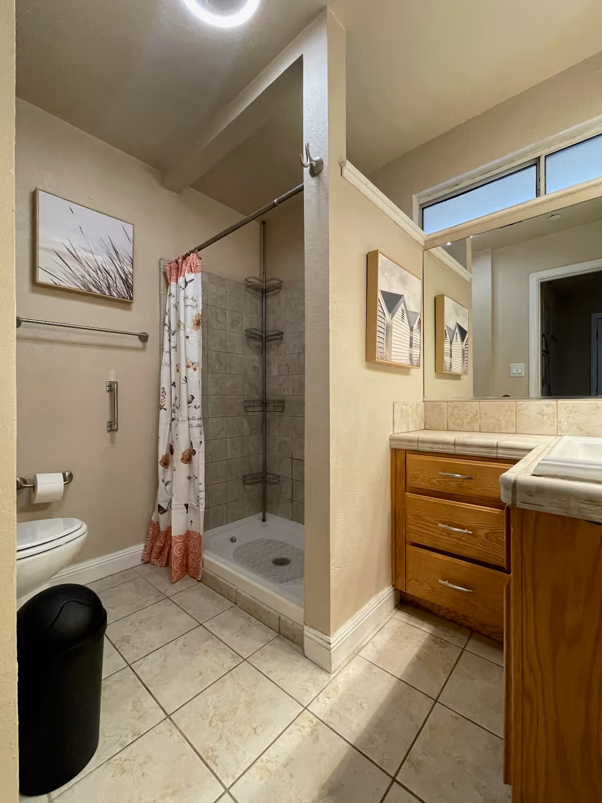 A bathroom with beige tiled floor and walls. There is a toilet with a black trash bin beside it, a shower with a floral shower curtain, and a vanity with wooden drawers and a tiled countertop. A large mirror is mounted above the vanity, and two framed pictures hang on the walls.