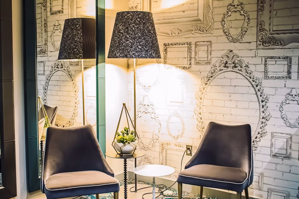 A cozy seating area with two gray upholstered chairs, a small round white table between them, and a tall floor lamp with a black patterned lampshade. The wall behind features a white brick design with decorative empty picture frames in various shapes and sizes. A large mirror is positioned on the left side, reflecting part of the scene.